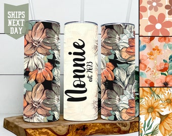 Könnte beinhalten: Drei Edelstahlbecher mit unterschiedlichen Designs. Der erste Becher hat ein Blumenmuster in Orange-, Rosa- und Grüntönen. Der zweite Becher ist weiß mit schwarzem Text, der "Mommie est 2023" lautet. Der dritte Becher hat das gleiche Blumenmuster wie der erste Becher.