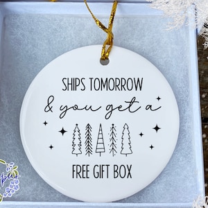 May include: A white ceramic Christmas ornament with black text that reads "SHIPS TOMORROW & you get a FREE GIFT BOX". The ornament has a gold ribbon for hanging and is inside a white gift box.