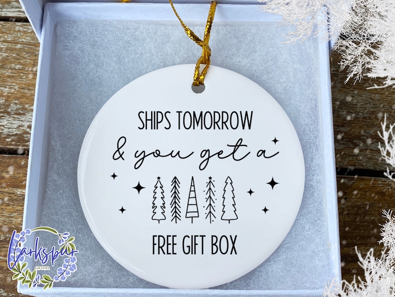 May include: A white ceramic Christmas ornament with black text that reads "SHIPS TOMORROW & you get a FREE GIFT BOX". The ornament has a gold ribbon for hanging and is inside a white gift box.