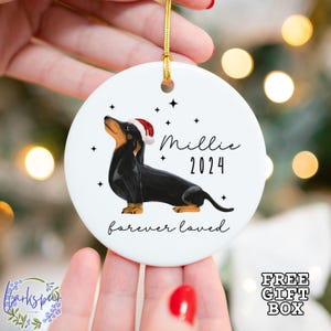K&ouml;nnte beinhalten: Wei&szlig;er Keramik-Weihnachtsbaumschmuck mit einem schwarzen Dackel, der einen roten Weihnachtsmann-Hut tr&auml;gt. Der Anh&auml;nger hat schwarze Sterne und den Text "Millie 2024 forever loved".