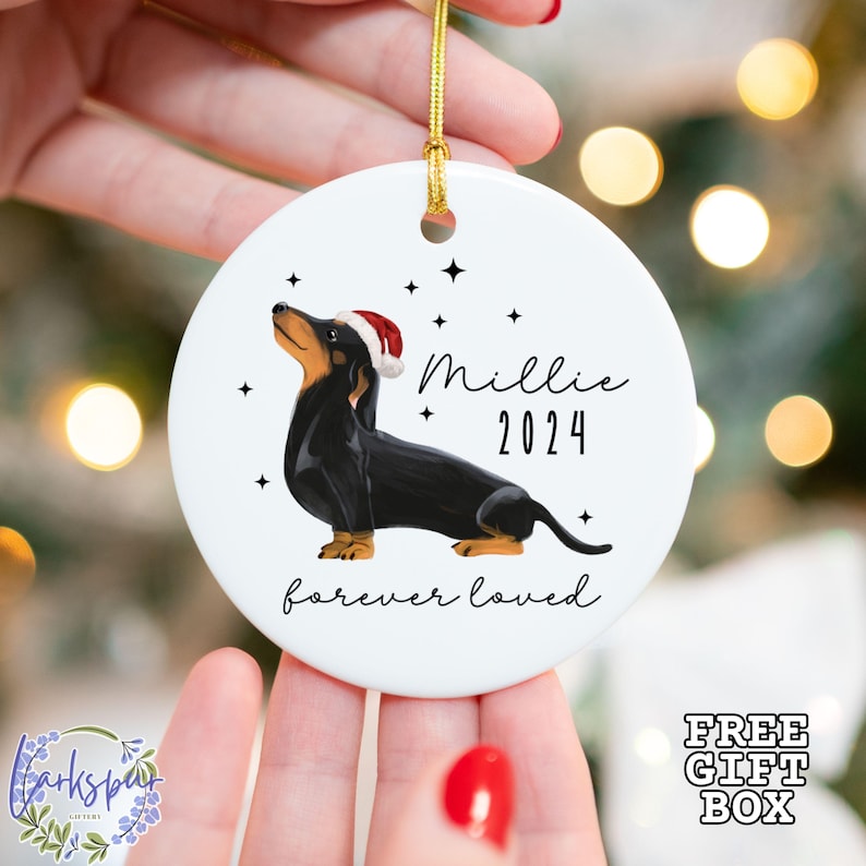 K&ouml;nnte beinhalten: Wei&szlig;er Keramik-Weihnachtsbaumschmuck mit einem schwarzen Dackel, der einen roten Weihnachtsmann-Hut tr&auml;gt. Der Anh&auml;nger hat schwarze Sterne und den Text "Millie 2024 forever loved".
