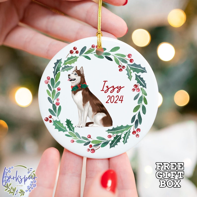 May include: A round white ceramic ornament featuring a brown and white husky dog illustration, surrounded by a green holly wreath with red berries. The name "Izzy" and the year "2024" are written in red script. A gold string is attached for hanging.