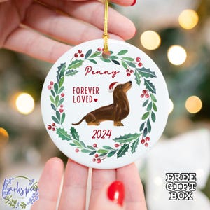 K&ouml;nnte beinhalten: Wei&szlig;er Keramik-Weihnachtsbaumschmuck mit einem gr&uuml;nen Stechpalmenkranz, roten Beeren und einem braunen Dackel, der einen Weihnachtsmann-Hut tr&auml;gt. Der Anh&auml;nger tr&auml;gt die Aufschrift "Penny Forever Loved" und "2024".
