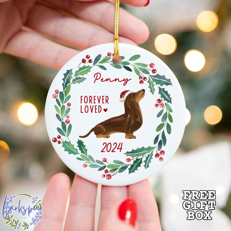 K&ouml;nnte beinhalten: Wei&szlig;er Keramik-Weihnachtsbaumschmuck mit einem gr&uuml;nen Stechpalmenkranz, roten Beeren und einem braunen Dackel, der einen Weihnachtsmann-Hut tr&auml;gt. Der Anh&auml;nger tr&auml;gt die Aufschrift "Penny Forever Loved" und "2024".