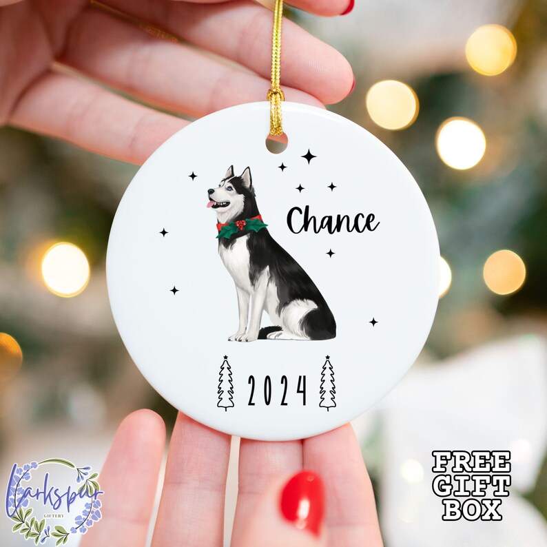 May include: White ceramic Christmas ornament featuring a black and white husky dog wearing a festive red and green collar. The ornament has the name "Chance" and the year "2024" printed on it, with star and tree accents.