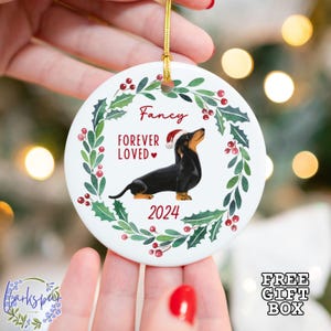 K&ouml;nnte beinhalten: Wei&szlig;er Keramik-Weihnachtsbaumschmuck mit einem gr&uuml;nen Stechpalmenkranz, roten Beeren und einem schwarz-braunen Dackel, der eine Weihnachtsm&uuml;tze tr&auml;gt. Der Schmuck ist mit dem Namen "Fancy" und dem Text "Forever Loved" mit einem Herzen personalisiert. Das Jahr "2024" ist ebenfalls enthalten.