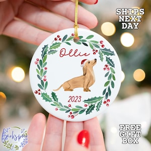 May include: White ceramic Christmas ornament with a green holly wreath, red berries, and a dachshund wearing a Santa hat. The ornament says "Ollie" and "2023".