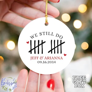 May include: White ceramic ornament with the text "We Still Do" and a tally mark graphic. The ornament also includes the names "Jeff & Arianna" and the date "09.16.2014".