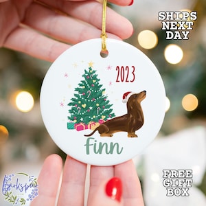 May include: White ceramic Christmas ornament with a brown dachshund wearing a Santa hat in front of a green Christmas tree with red and white ornaments. The ornament has the text "2023" and "Finn" on it.