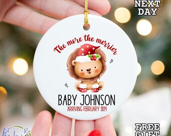 Könnte beinhalten: Weißer Keramik-Weihnachtsbaumschmuck mit einem Löwen, der einen Weihnachtsmann-Hut trägt, und dem Text "The more the merrier" und "Baby Johnson Arriving February 2024".