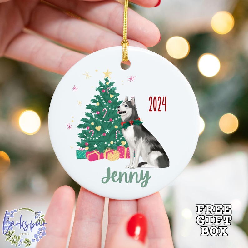 May include: White ceramic Christmas ornament featuring a Siberian Husky with a Christmas tree, gifts, and the year 2024. The name "Jenny" is printed below the dog. A gold string is attached for hanging.