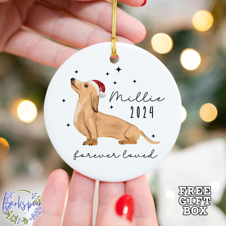 K&ouml;nnte beinhalten: Wei&szlig;er Keramik-Weihnachtsbaumschmuck mit einem braunen und braunen Dackel, der einen roten Weihnachtsmann-Hut tr&auml;gt. Der Anh&auml;nger hat den Text "Millie 2024 forever loved" darauf.