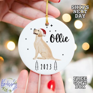 May include: White ceramic Christmas ornament featuring a yellow Labrador Retriever wearing a red Santa hat. The dog is sitting and looking to the left. The ornament also includes the text "Ollie" and "2023" with two stylized Christmas trees.