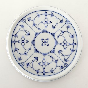 Könnte beinhalten: Eine runde Untertasse aus weißem Porzellan mit einem blauen Unterglasur-Muster. Das Design besteht aus einem zentralen kreisförmigen Motiv, umgeben von vier symmetrischen Abschnitten, die jeweils zarte Blumenranken und eine vierblättrige Blüte enthalten.