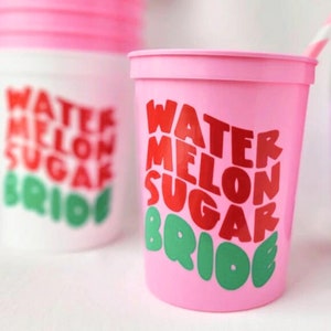 Puede incluir: Un vaso de plástico rosa con el texto "Watermelon Sugar Bride" en letras rojas y verdes. El vaso tiene una pajita.