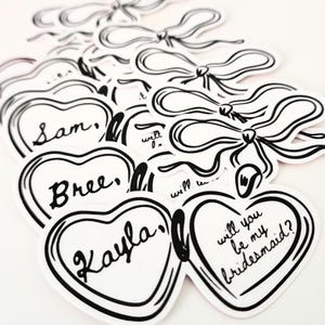 May include: A set of six white heart-shaped stickers with black outlines and black text. The stickers say "Will you be my bridesmaid?" and include the names "Sam", "Bree", and "Kayla".