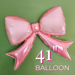 May include: A large pink bow-shaped balloon, measuring 104 cm across, against a green background. The balloon has a glossy finish and is designed to resemble a tied bow. The word "BALLOON" is printed below.