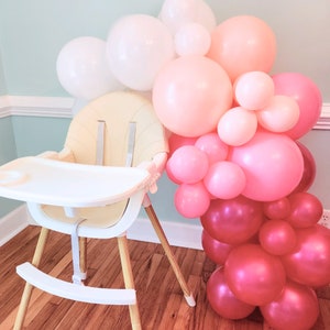May include: A white high chair with a beige padded seat and a white tray. The chair is in front of a wall decorated with a balloon arch made of pink, peach, and white balloons.
