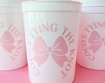 May include: White plastic cups with pink text and a pink bow design that says "Coco's Tying The Knot".