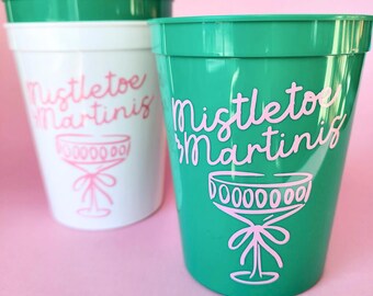 以下が含まれることがあります： 緑と白の外側のプラスチック製のカップが2つ。カップにはピンクの文字で「Mistletoe Martinis」と書いてあり、ピンクのマーティーニグラスのイラストが描かれています。