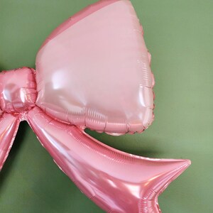 May include: Close-up of a large, pink bow-shaped balloon. The balloon has a shiny, metallic finish and is composed of several connected sections. The bow is a light pink color with a darker pink outline, and is against a green background.