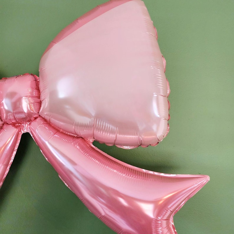 May include: Close-up of a large, pink bow-shaped balloon. The balloon has a shiny, metallic finish and is composed of several connected sections. The bow is a light pink color with a darker pink outline, and is against a green background.