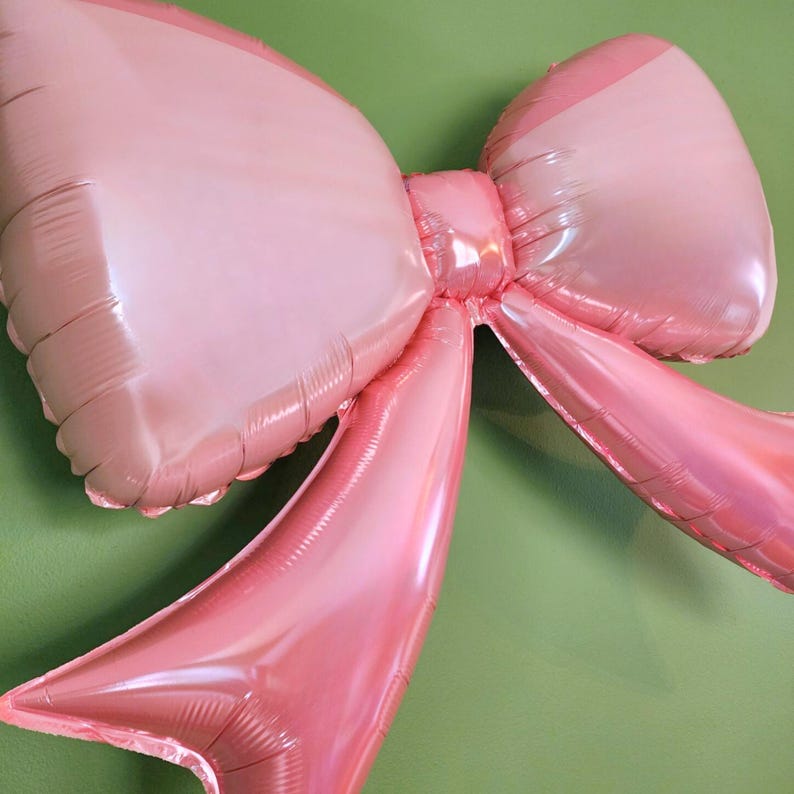 May include: A large, pink bow-shaped balloon against a green background. The balloon has a shiny, metallic finish and is inflated, with the bow's loops and tails clearly defined. The color is a soft, pastel pink.