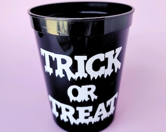 Pode incluir: Copo de plástico preto com o texto "Trick or Treat" em branco em uma fonte de gotejamento.