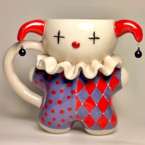 May include: A ceramic mug shaped like a jester with red and blue accents. The mug has a white face with black cross eyes and a red nose. The mug has a white ruffle collar and a handle that is shaped like a jester's arm.