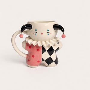 May include: A whimsical ceramic mug shaped like a clown. The mug features a white face with black horns, blue and red accents, and a ruffled collar. The body is split between a pink polka-dot pattern and a black and white checkerboard design.