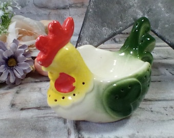 May include: A ceramic chicken egg holder with a yellow body, red comb, and green wings. The holder has a white interior for holding eggs.