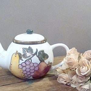 Puede incluir: Tetera de cerámica blanca con tapa, con un diseño pintado a mano de frutas, incluyendo una pera, uvas y una manzana. La tetera tiene un borde dorado y un pomo verde. Un ramo de rosas rosas claras descansa junto a ella.