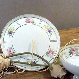 Puede incluir: Un juego de dos platos de porcelana vintage con un diseño floral. El plato más grande tiene un centro blanco con un borde dorado y un patrón floral alrededor del borde. El plato más pequeño tiene un diseño similar pero es más pequeño.