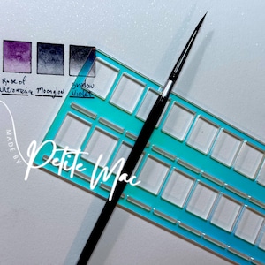 Op de afbeelding: Een turquoise en transparant aquarelpalet met een zwarte penseel. Het palet heeft meerdere lege vierkante putjes. Drie aquarelkleurstalen worden weergegeven, met de labels "Rose of Ultramarine", "Moonglow" en "Shadow Violet". De tekst "Petite Mac" is cursief geschreven.