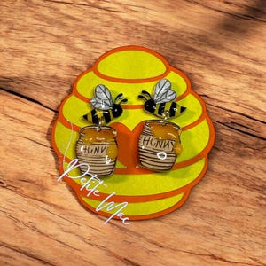 May include: A pair of honeybee earrings with a honey pot charm. The earrings are on a yellow honeycomb-shaped display card with the text "Petite Mac".