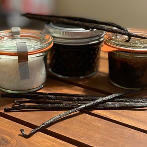May include: Three glass jars with orange-rimmed lids and a metal clasp, filled with vanilla products, sit on a wooden surface. One jar contains white crystals, another dark vanilla beans, and the third a dark liquid. Several vanilla beans are scattered around.