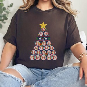 Op de afbeelding: Een bruin t-shirt met een kerstboomontwerp gemaakt van honkballen. De boom is bekroond met een gouden ster en versierd met feestelijke lichtjes en cadeaus. Het t-shirt wordt door een persoon gedragen.