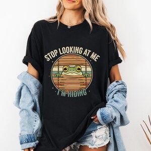 Puede incluir: Camiseta negra con un diseño de rana de dibujos animados. La rana está mirando a través de persianas, con el texto "STOP LOOKING AT ME I'M HIDING" en un círculo. La rana es verde y las persianas son marrones.