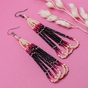 May include: A pair of handmade beaded earrings with a pink background. The earrings feature a combination of black, cream, and pink beads. The design includes a fringe of black beads and a beaded loop at the bottom. The earrings are accented with gold-colored beads.