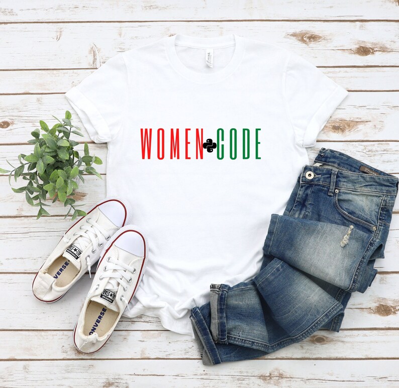 Peut inclure: T-shirt blanc avec le texte "WOMEN+CODE" en rouge, noir et vert. Le t-shirt est pos&eacute; sur une surface en bois avec une paire de baskets blanches et un jean bleu.