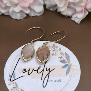 Op de afbeelding: Goudkleurige oorbellen met druppelvormige, lichtbruine en beige edelstenen. De oorbellen worden gepresenteerd op een witte kaart met het woord "Lovely" en de tekst "by Zoe". Decoratieve bloemen staan op de achtergrond.