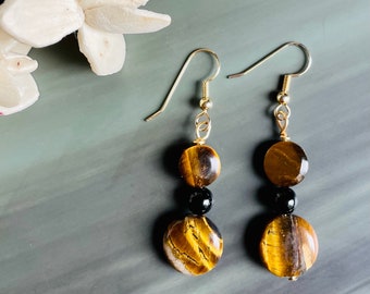 Op de afbeelding: Gouden oorhangers met tijgeroog en zwarte kralen. De oorbellen hebben een eenvoudig ontwerp met twee ronde tijgeroogkralen en een zwarte kraal ertussen.