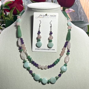May include: A necklace and earring set displayed on a mannequin. The necklace features a layered design with various gemstone beads in shades of green, pink, purple, and silver accents. The earrings mirror the necklace's color scheme. The tag reads "jasper rain design studio".