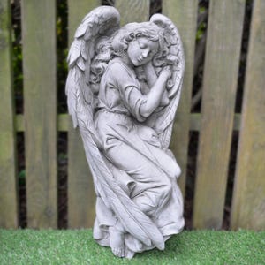 May include: A grey stone sculpture of an angel with wings, sleeping with its head resting on its hand. The angel is sitting with its legs crossed and its wings are spread out behind it.