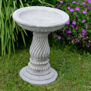 May include: A grey concrete bird bath with a round basin and a decorative pedestal. The pedestal has a twisted design and a beaded base. The bird bath is on a grassy lawn, with green foliage and purple flowers in the background.