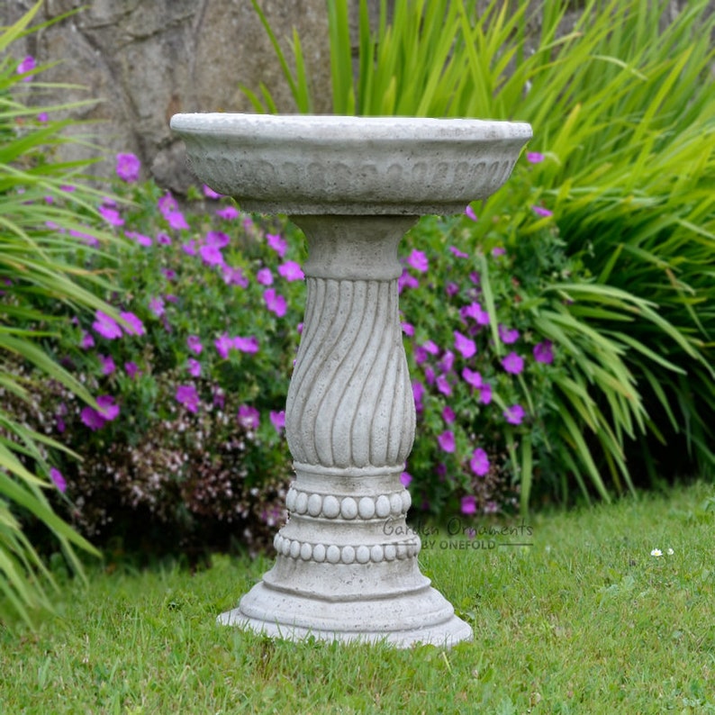 Op de afbeelding: Een grijs betonnen vogelbad met een decoratieve, wervelende voet. Het vogelbad heeft een brede, ondiepe kom met een gedetailleerde rand. De basis is rond en staat op een grasveld, met paarse bloemen en groene bladeren op de achtergrond.