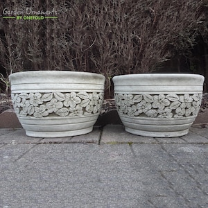 Puede incluir: Dos macetas de flores de hormigón blanco con un diseño floral. Las macetas son de diferentes tamaños y están sentadas en un patio gris.