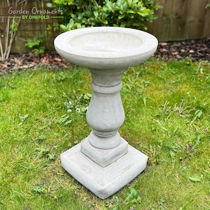 Puede incluir: Un bebedero para pájaros de hormigón gris con un cuenco redondo y una base de pedestal. El bebedero para pájaros está situado en un césped verde.