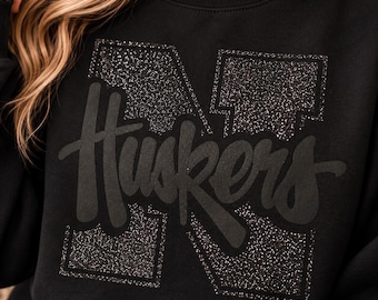 Pode incluir: Uma camisola preta de gola redonda com uma letra N grande e brilhante no fundo. Sobreposta à letra está a palavra Huskers escrita num tipo de letra escuro, mate e cursivo. O design está centrado no peito da peça de vestuário.
