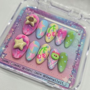May include: A set of ten press-on nails in a clear plastic case. The nails feature various designs, including stars, cherries, and abstract patterns in pink, blue, yellow, and green. The case has text that reads "HOME", "SHOP", "CART", and "INSTA: @Veenailsit_".
